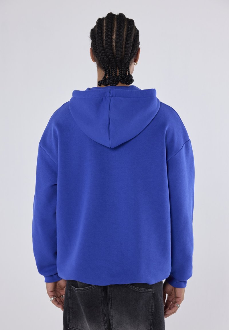 Person mit geflochtenem Haar, der einen königsblauen Kapuzenpullover und ausgewaschene schwarze Jeans trägt, steht vor einem schlichten hellen Hintergrund mit dem Rücken zugewandt.