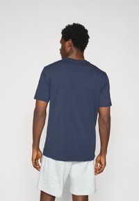 Marineblaue Baumwoll-T- shirt mit Rundhalsausschnitt und kurzen Ärmeln, von hinten betrachtet, kombiniert mit hellgrauen Shorts.