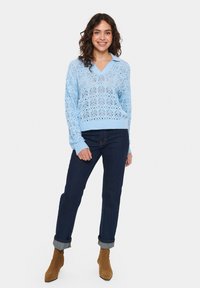 Gaiši zils adīts džemperis ar V-veida kakla izgriezumu un mežģīņu rakstiem, apvienots ar tumšiem džinsa biksēm. Models valkā brūnas potītes zābaciņus.