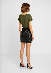 Camisa de manga corta verde oliva combinada con una mini falda negra de lentejuelas. Tacones altos negros completan el atuendo. Tela suave y lentejuelas brillantes visibles.