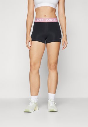 Crne atletske kratke hlače s roza Nike Pro pojasom, s uskim fitom i glatkom teksturom tkanine. Nosi se s laganim tenisicama i bijelim čarapama.