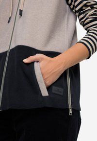 Giacca in pile con colori grigio e blu navy, chiusura con zip, maniche a righe e tasche laterali. Include una patch in pelle con logo sulla parte anteriore.