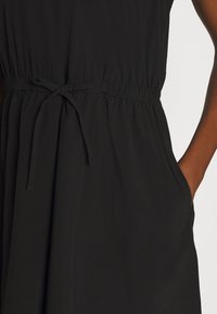 Robe noire avec une taille élastique resserrée, présentant un détail noué, des poches latérales et un tissu doux et texturé.