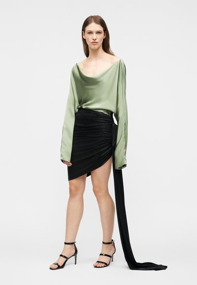 Γυναίκα που φοράει μία ανοιχτόχρωμη πράσινη σατέν μπλούζα και μια μαύρη ασύμμετρη φούστα με μία μακριά μαντήλα που κρέμεται, σε συνδυασμό με μαύρα σανδάλια υψηλού τακουνιού.