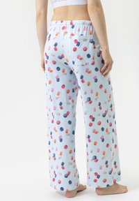 Vrouw staat blootvoets in lichtblauwe pyjamabroek met een multicolor macaron print en een witte crop top, gezien van achteren.