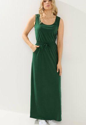 Kobieta ubrana w długą, bezrękawnikową sukienkę maxi w ciemnozielonym kolorze z elastycznym pasem i kieszeniami, stojąca na jasnym, jednolitym tle.