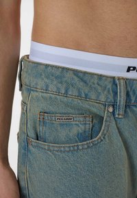 Light blue denim jeans with a textured surface, five-pocket design, and orange stitching. White elastic waistband with black logo visible.