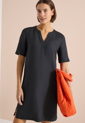 Mujer con vestido negro hasta la rodilla y mangas cortas sosteniendo una chaqueta acolchada naranja brillante sobre un fondo beige.