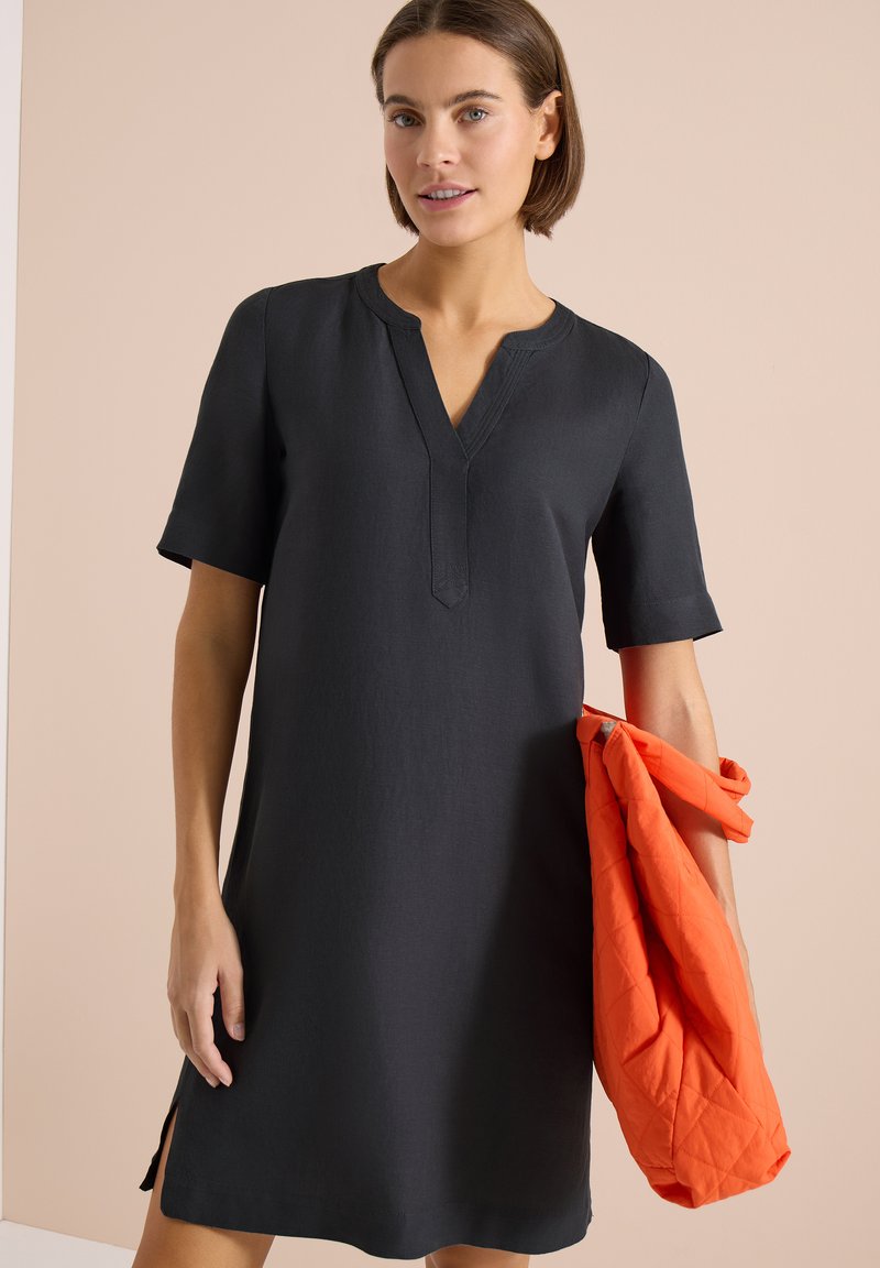 Femme portant une robe noire arrivant au genou avec des manches courtes, tenant une veste matelassée orange vif sur un fond beige.
