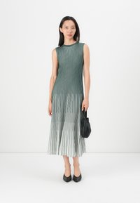 Sleeveless pleated dress in dark green and cream, featuring vertical stripes. Model holds a small black handbag, wearing pointed-toe pumps.
