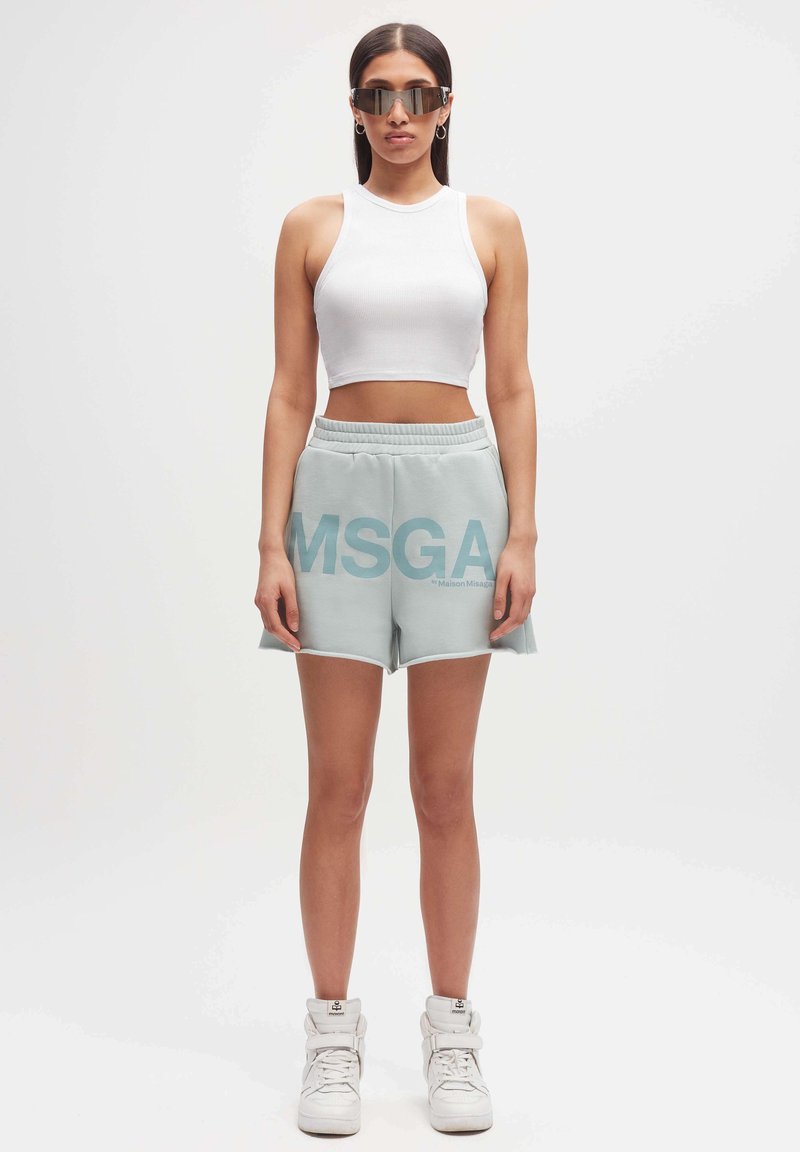 Lichtblauwe katoenen shorts met grote "MSGA" opdruk, elastische tailleband, gecombineerd met een witte geribbelde crop top en witte hoge sneakers.