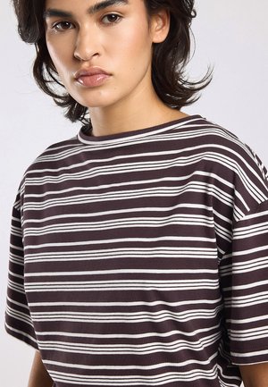 Jong persoon met middellang donker haar, draagt een kortarmig bruin-wit horizontaal gestreept shirt, neutrale gezichtsuitdrukking, effen achtergrond.