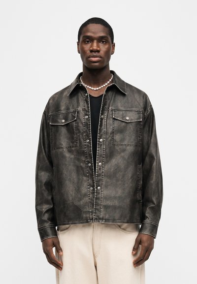 Νεαρός άνδρας με σκούρο καφέ παλιό δερμάτινο μπουφάν πάνω από μαύρο πουκάμισο, μπεζ παντελόνι και ασημένιο αλυσιδάκι, στέκεται μπροστά σε λευκό φόντο.