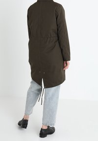 Abrigo largo de color verde oliva con cintura ajustada, que presenta un dobladillo redondeado y un detalle de cordón en la parte de atrás, combinado con vaqueros de color azul claro y zapatos negros.