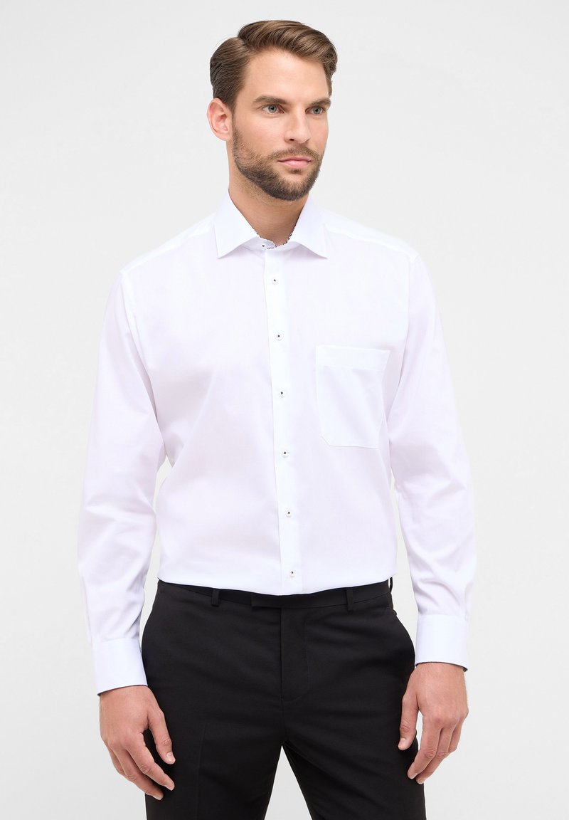 Camicia bianca a maniche lunghe con bottone frontale, tasca singola e bottoni neri. Tessuto liscio e vestibilità su misura, abbinata a pantaloni neri.