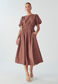 Robe midi marron à pois, avec des manches bouffantes, une taille élastique et des poches latérales. Associée à des talons blancs à lanières.
