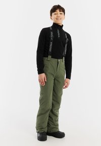 Groene sneeuwbroek met verstelbare zwarte bretels met "PROTEST" merk. Zacht materiaal, ruime pasvorm, gedragen met een zwart langmouwtop en laarzen.