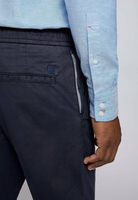 Ljust blå, kragklädd skjorta med en mjuk textur. Mörkblå byxor med en sidofickan med dragkedja och en liten accentlogotyp.