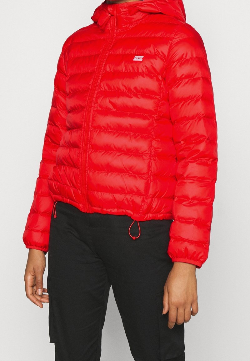 Chaqueta acolchada roja con cremallera frontal, capucha y cordones ajustables en el dobladillo y los puños. Textura suave con patrones horizontales cosidos.