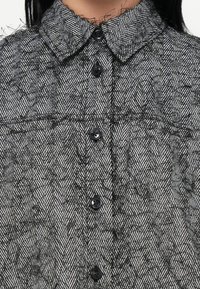 Close-up van een zwart-wit gestreept shirt met een hoge kraag, uitgevoerd in een visgraatpatroon en zwarte knopen aan de voorkant.