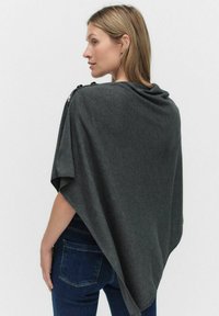 Szary dzianinowy poncz z otwartym krojem, z dekoltem w kształcie draperii i guzikami przy ramieniu. Miękka tekstura, noszone na ciemnych dżinsach.