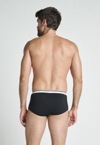 Svarta bomullstrosor med en vit märkesmidja, figursydd design och slät yta, sedd bakifrån.