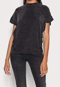 Sort kortærmet top med metalisk glans, voluminøse sideaccenter og høj hals, parret med mørke jeans. Glat tekstur.