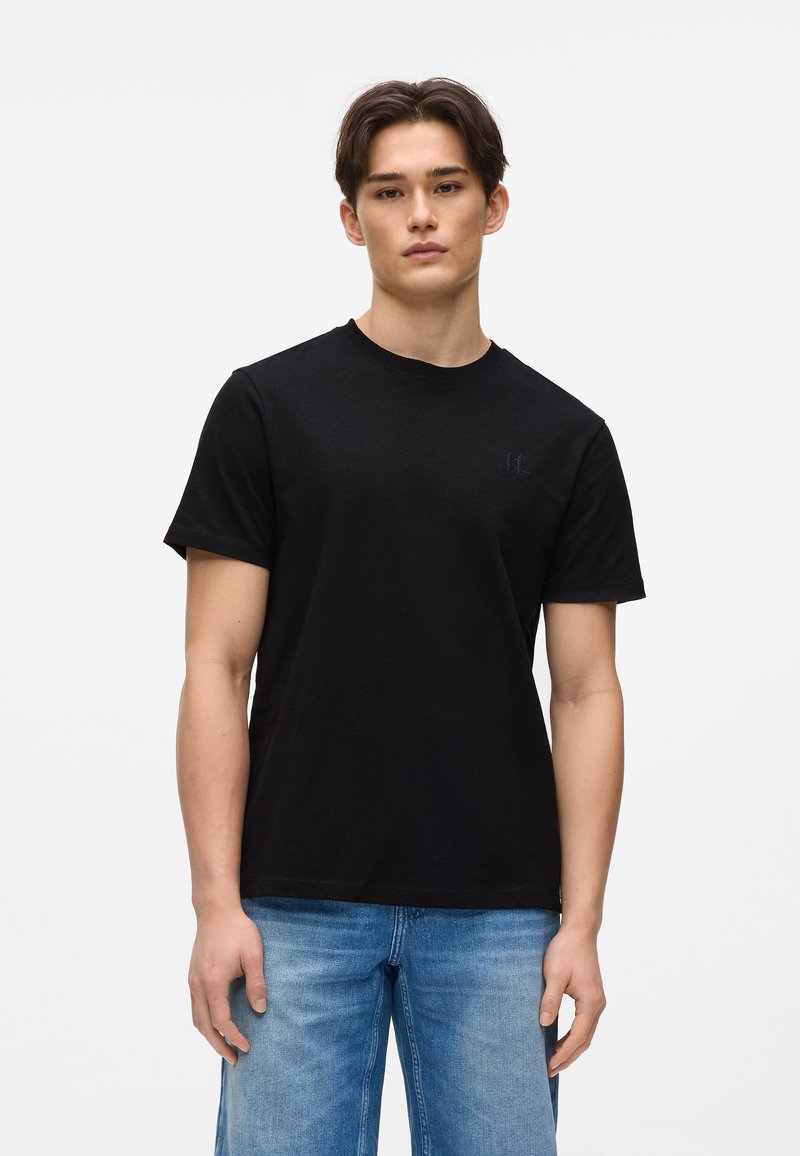 T-shirt en coton noir à manches courtes et col rond. Présente un logo discret sur la poitrine. Associé à un jean bleu clair.