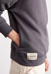 Pulover gri din bumbac cu guler tricotat, buzunar lateral și etichetă cu inscripția "STUDIO EDITION". Material texturat cu manșete canelate.