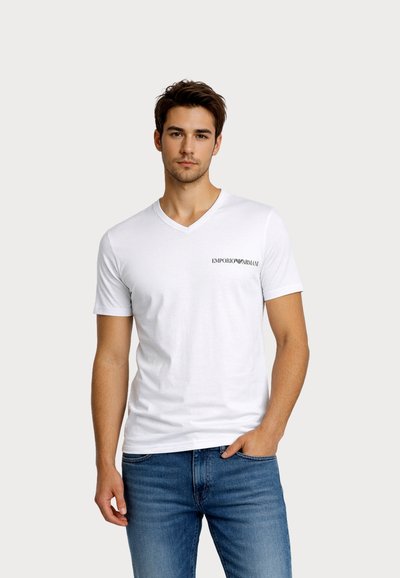 Νεαρός άνδρας που φοράει λευκό T-shirt V-neck Emporio Armani και μπλε τζιν, στέκεται με το ένα χέρι στην τσέπη μπροστά σε απλό φόντο.