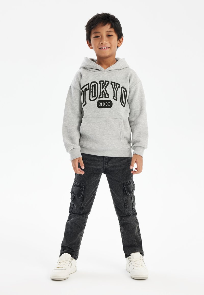 Sudadera gris con el texto "TOKYO" y "MOOD" debajo, combinada con pantalones cargo negros con bolsillos laterales y zapatillas blancas. Diseño sencillo, estilo casual.