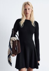 Schwarzes geripptes Strickkleid mit schwingendem Rock und langen Ärmeln. Das Model hält eine braune Wildledertasche, die mit einem gemusterten Seidenschal akzentuiert ist.