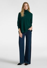 Maglione verde a collo alto con maniche lunghe, fessure laterali, abbinato a jeans in denim scuro a gamba larga. Colore uniforme, trama liscia e vestibilità casual.