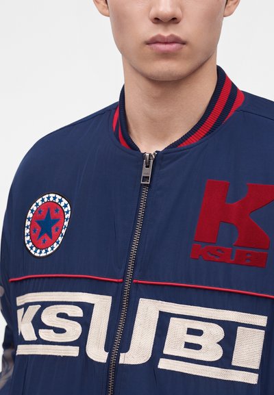 Jeune personne portant une veste zippée bleu marine avec des logos Ksubi rouges et blancs et un écusson en forme d’étoile sur la poitrine.