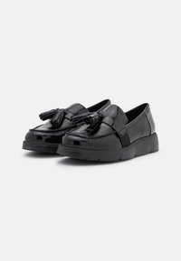 Mocassins en cuir verni noir avec une semelle légèrement surélevée et texturée, ornées de franges décoratives à l'avant.