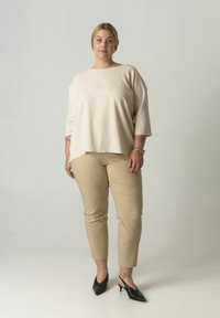 Beiges, locker sitzendes Oberteil mit Dreiviertelärmeln, kombiniert mit engen beigen Hosen und schwarzen spitzen Absatzschuhen. Glatte Stoffstruktur.
