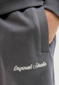 Pelēka sporta krekla audums ar ribota faktūra, ar sānu kabatu un baltu izšuvumu "Originals Studio" gar vīlīti.