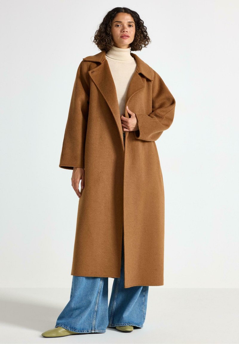 Cappotto lungo in lana color cammello con colletto a rever, vestibilità oversize e tasche laterali; indossato sopra un dolcevita beige e jeans a gamba larga.