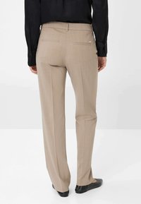 Beige maßgeschneiderte Hose mit geradem Schnitt, ausgestattet mit einer glatten Textur und zwei Gesäßtaschen. Dazu schwarze Schuhe.