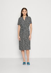 Svart klänning med vit blommig tryck, korta ärmar och knytmidja. Platta sandaler med teal-färgade klackar. Slät tyg och knä-lång design.