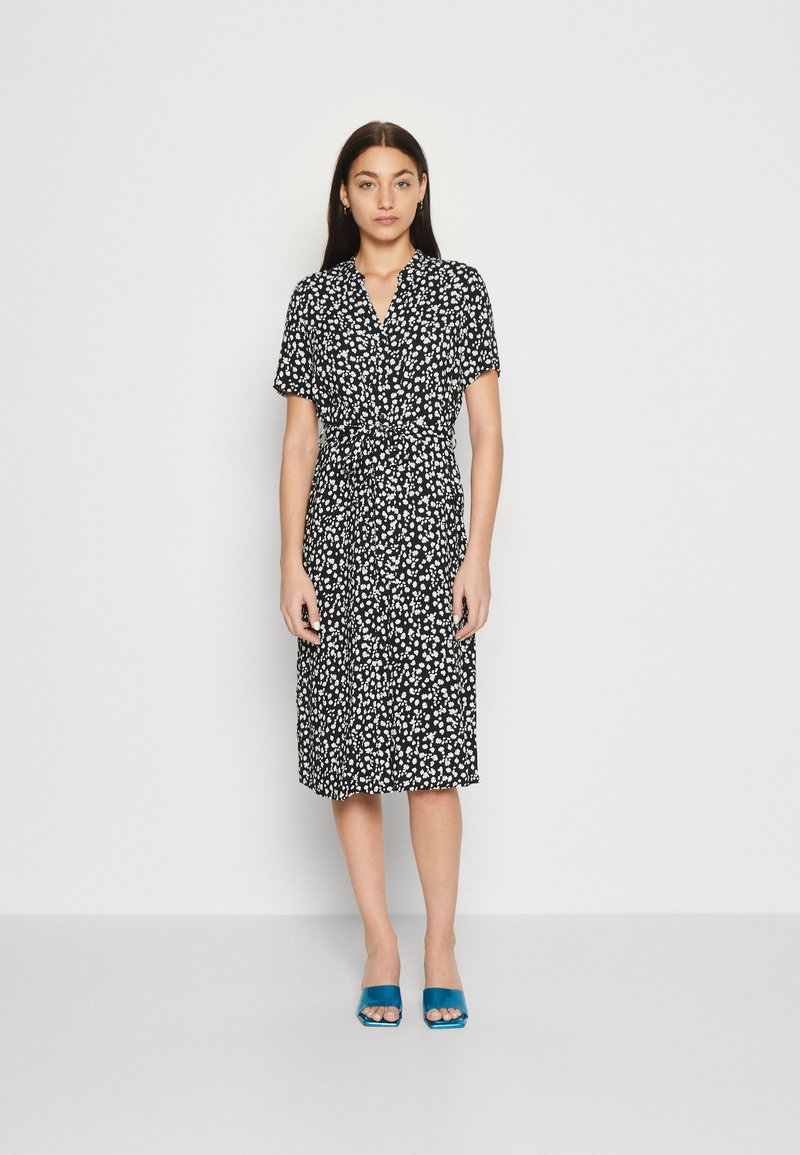 Svart klänning med vit blommig tryck, korta ärmar och knytmidja. Platta sandaler med teal-färgade klackar. Slät tyg och knä-lång design.