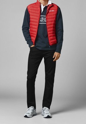 Mężczyzna ubrany w czerwoną puchową kamizelkę na granatowej koszuli z logo, czarne spodnie i białe trampki, stojący z jedną ręką w kieszeni.