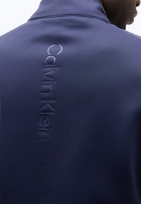 Άνδρας που φοράει μπλε σκούρο μπουφάν με κάθετα ανάγλυφο λογότυπο Calvin Klein στην πάνω πλάτη, στραμμένος ελαφρώς προς το πλάι από την κάμερα.