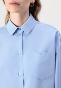 Camicia azzurra chiaro con colletto classico, due bottoni e taschino sul petto con logo ricamato "2NDDAY". Tessuto morbido.