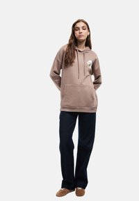 Brązowy hoodie z kangurzą kieszenią, kapturem ze sznurkiem i haftowanym wzorem. Połączony z ciemnoniebieskimi spodniami typu wide-leg i beżowymi butami slip-on.