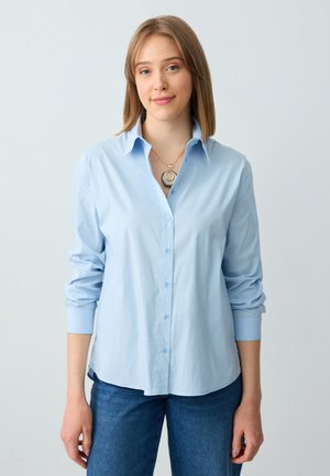 Young woman with straight blonde hair wearing a light blue button-up shirt, blue jeans, and a layered circular pendant necklace.