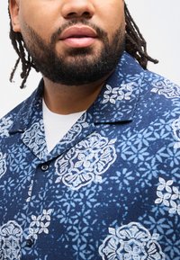 Άνδρας με γένια και σκουρόχρωμες κοτσίδες, φορώντας ένα μπλε πουκάμισο με σχέδια πάνω από ένα λευκό εσώρουχο, με το πρόσωπο στραμμένο ελαφρώς στο πλάι.