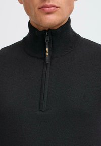 Svart stickad zip-up-tröja med ribbad hög krage och en partiell dragkedja med gulddetalj. Slät textur och figurnära design.