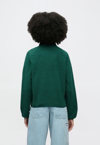 Grønn fleecejakke med høy krage, raglanermer og en avslappet passform, kombinert med lyseblå bukser med høyt liv.