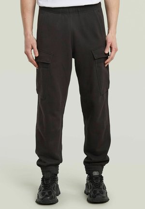 Black cargo jogger pants with elastic cuffs, side flap pockets, worn with black athletic shoes and a white tucked-in shirt.